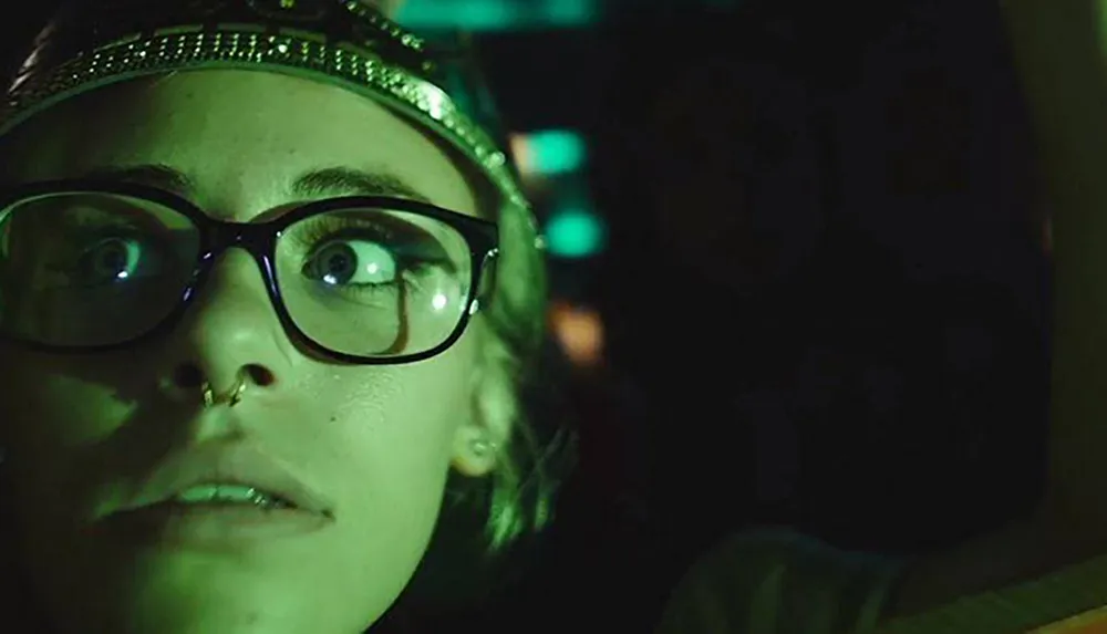 A person wearing glasses appears surprised or anxious in a dimly lit greenish environment
