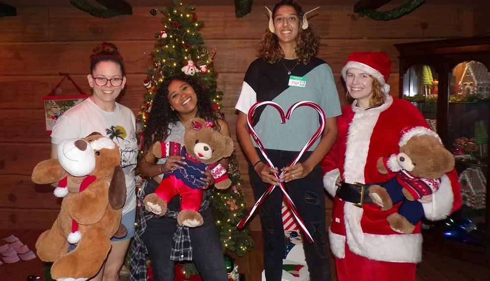 Four people are smiling and posing for a festive photo with plush teddy bears colorful decorations and a Christmas tree in the background