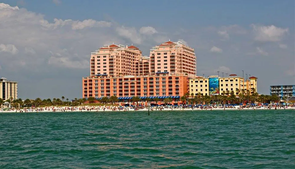 The image shows a vibrant beachfront lined with colorful buildings and a bustling beach crowd under a partly cloudy sky