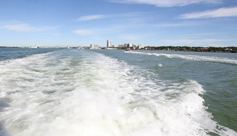 The image shows the wake of a boat with frothy waves looking back towards a coastline with buildings and a bridge under a clear blue sky