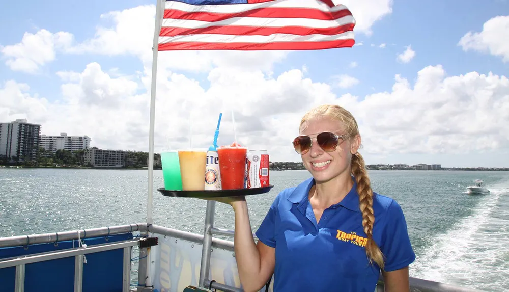 A smiling woman in sunglasses is holding a tray with colorful frozen drinks on a boat with the American flag and waterfront scenery in the background