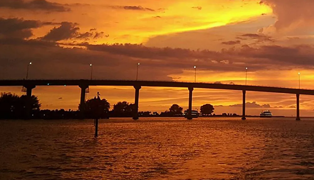 The image captures a serene sunset with vibrant orange and yellow hues in the sky silhouetting a bridge over a calm body of water