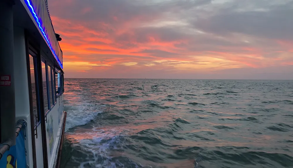 A breathtaking sunset paints the sky with hues of pink and orange as viewed from the side of a boat with blue lights on the ocean