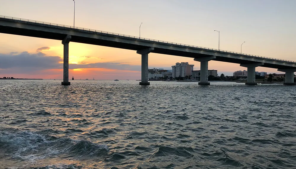 A long bridge spans across a body of water against the backdrop of a beautiful sunset with a silhouette of a lighthouse and buildings in the distance
