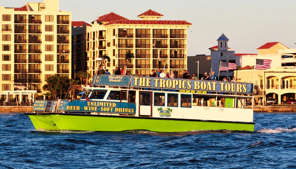 A colorful tour boat named THE TROPICS BOAT TOURS is cruising near the shore with passengers on board and American flags flying in the background
