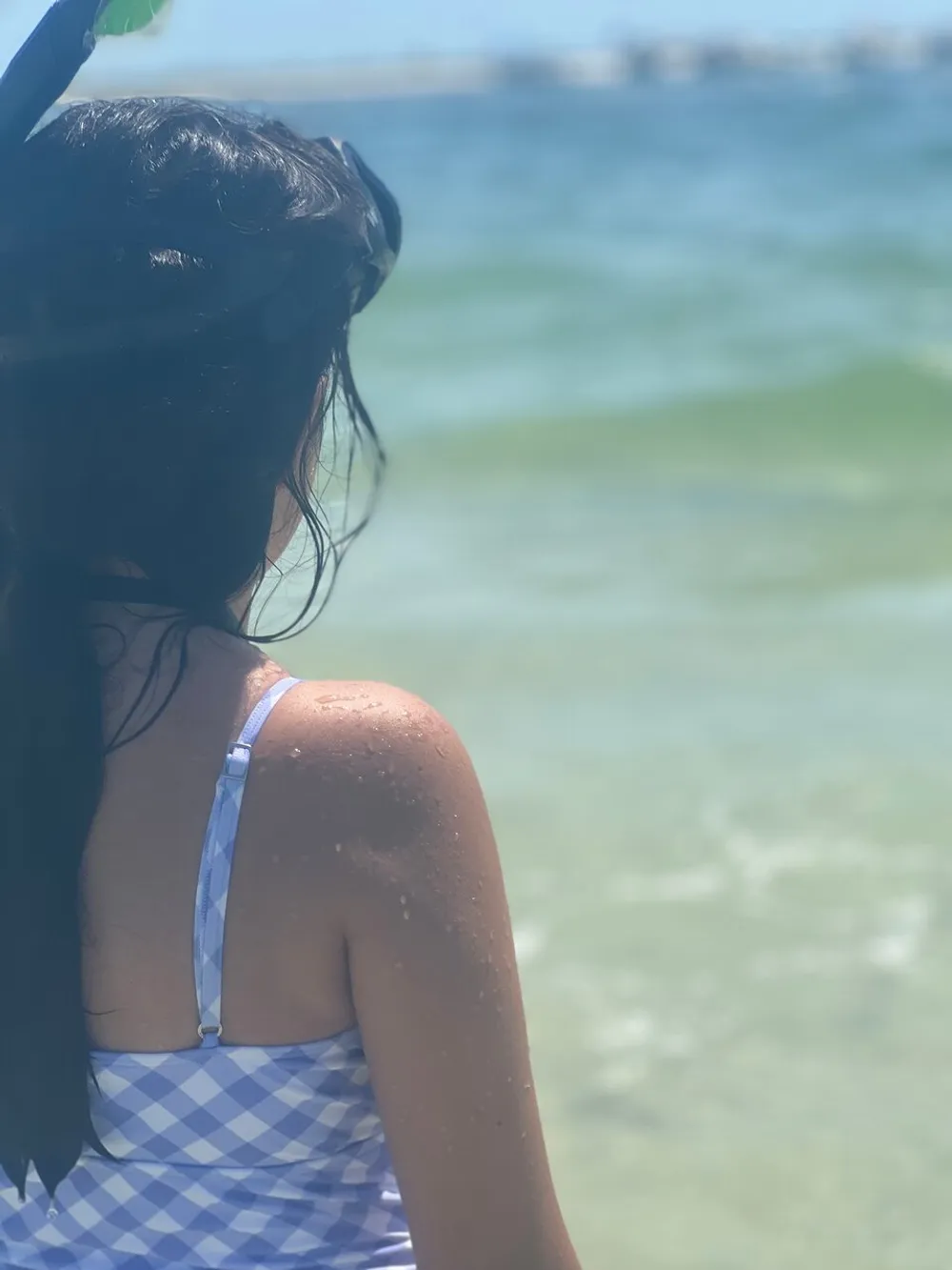 A person in a blue checkered swimsuit is gazing out at the sea with droplets of water on their skin suggesting they may have been enjoying a swim