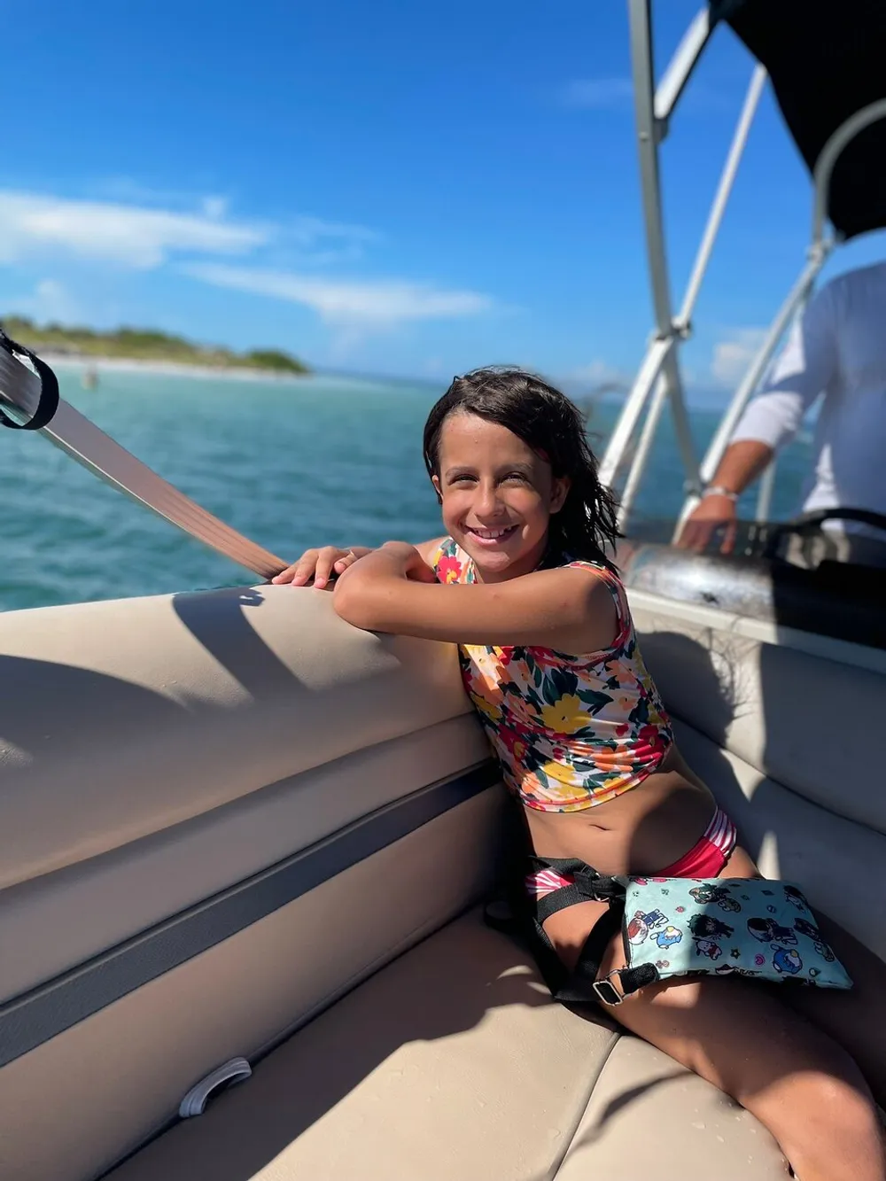 A smiling child in a colorful swimsuit is seated comfortably on a boat with a clear blue sky and water in the background