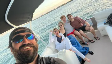 A group of five people are posing for a selfie on a boat, with the person at the front holding the camera and everyone appearing happy and relaxed, set against a backdrop of calm waters and a distant coastline under a clear sky.