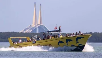 A yellow tour boat adorned with dolphin graphics speeds through the water with passengers on board, with a large cable-stayed bridge visible in the background.