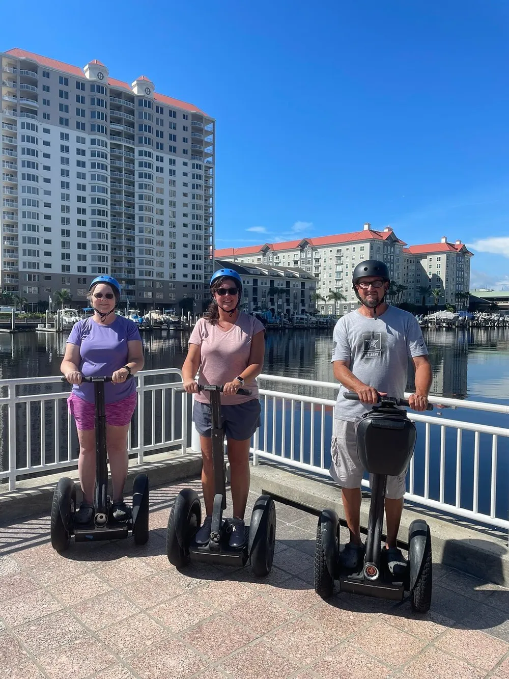 Three people wearing helmets are standing on Segways by a waterfront with buildings in the background on a sunny day