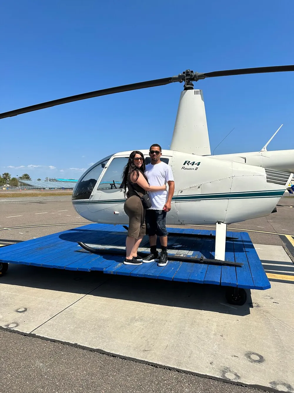 Two people are posing for a photo in front of a white R44 Raven II helicopter on a sunny day