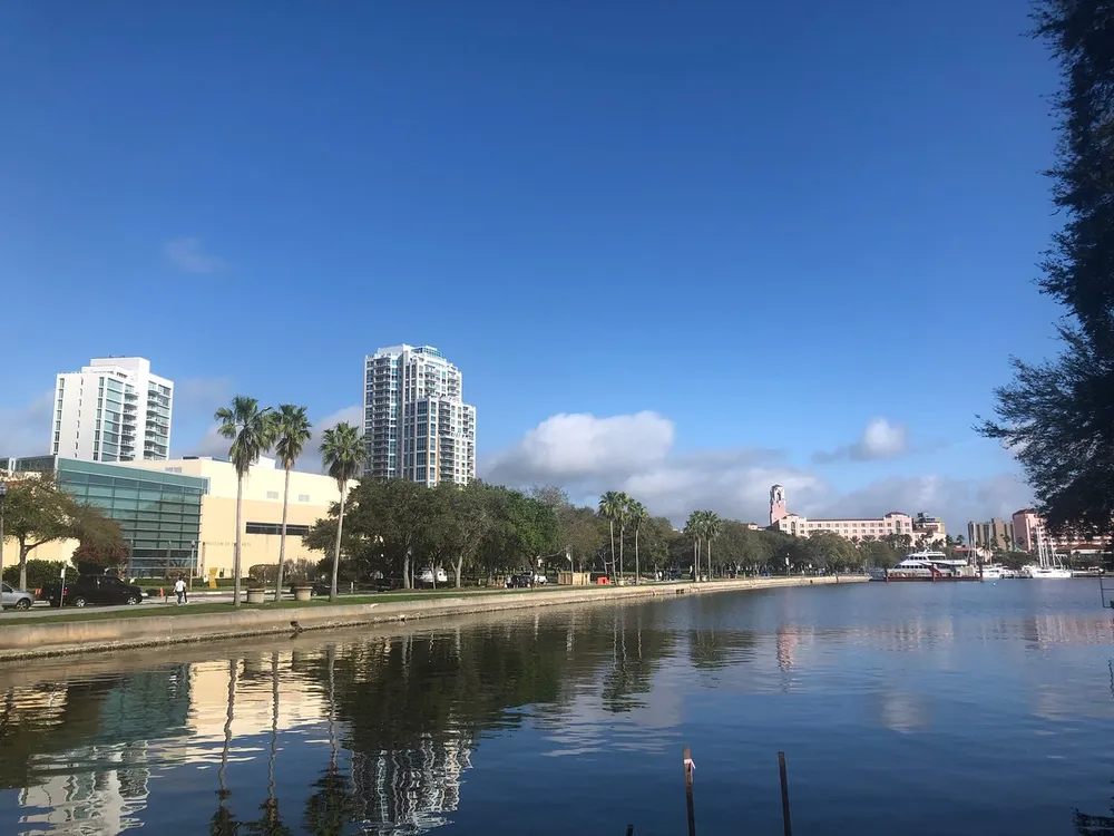 The image depicts a serene riverside scene with modern buildings palm trees and a clear blue sky reflecting in the calm waters of the river