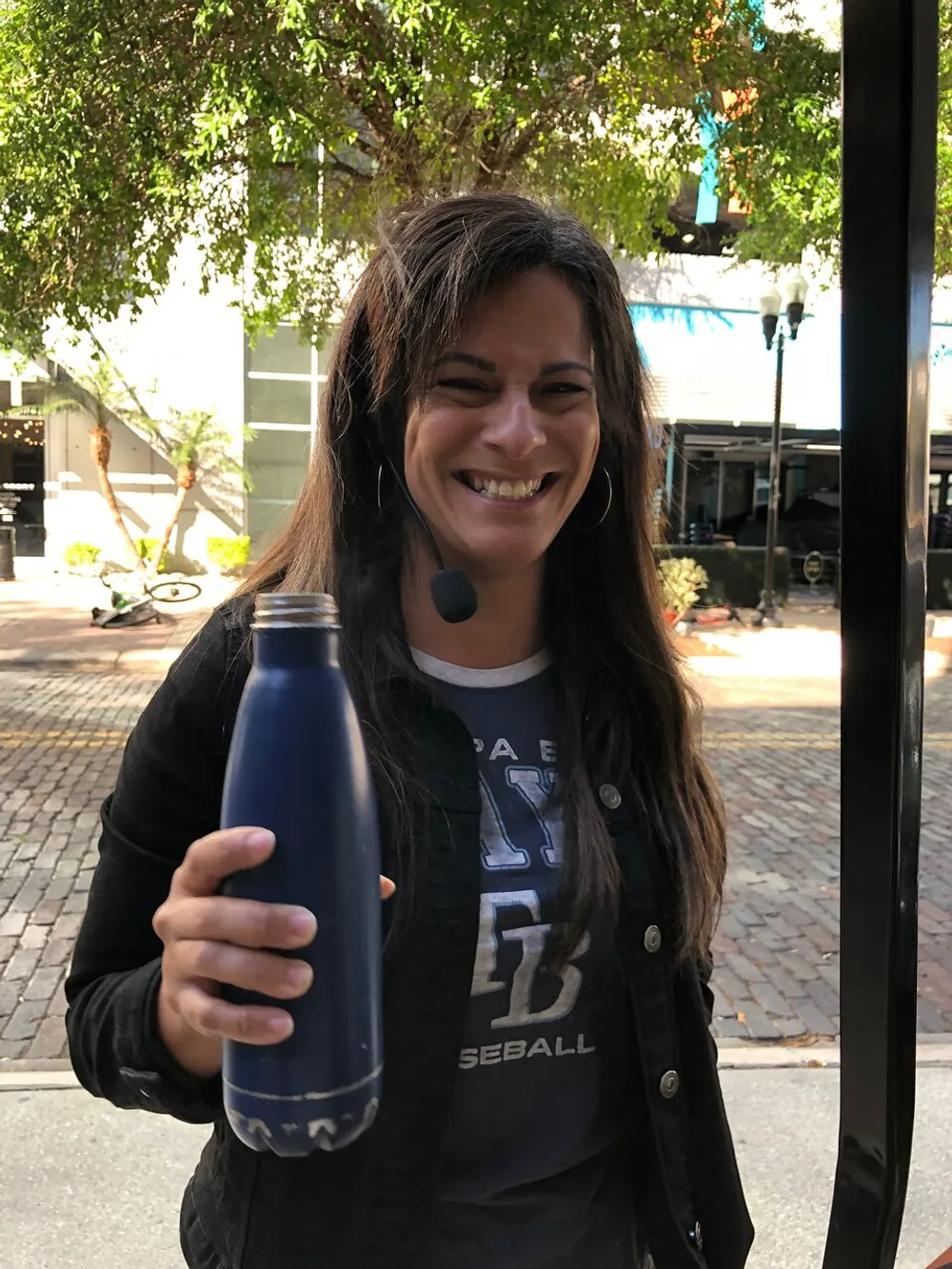 A smiling woman with a headset is holding a blue water bottle outdoors