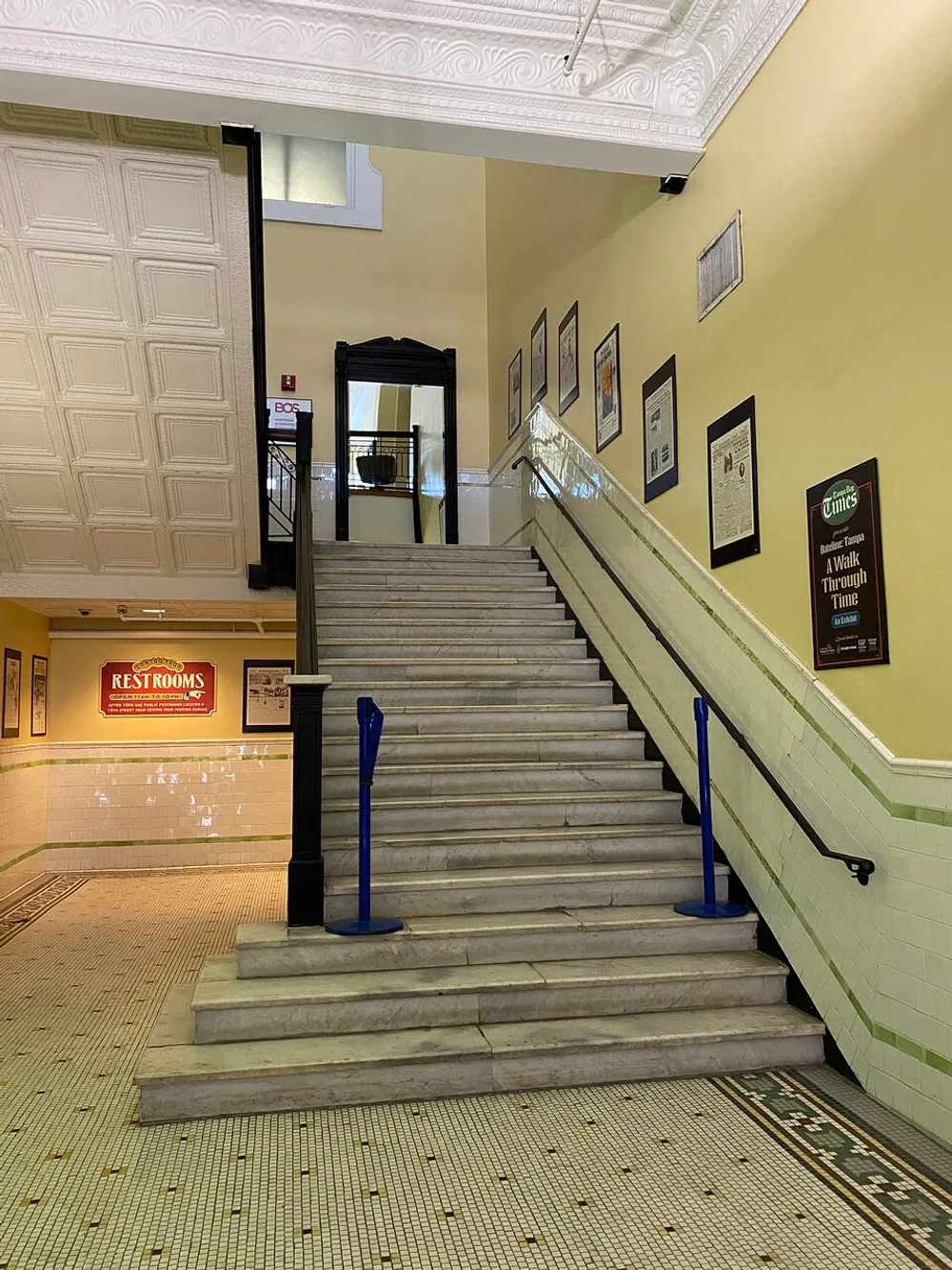 The photo shows an indoor staircase with decorative tiled floors framed pictures on the walls and signs indicating restrooms and an exhibition likely situated within a historic or public building