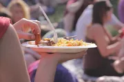A person is holding a plate of food with a fork, and there are other people in the blurry background, possibly at an outdoor event.