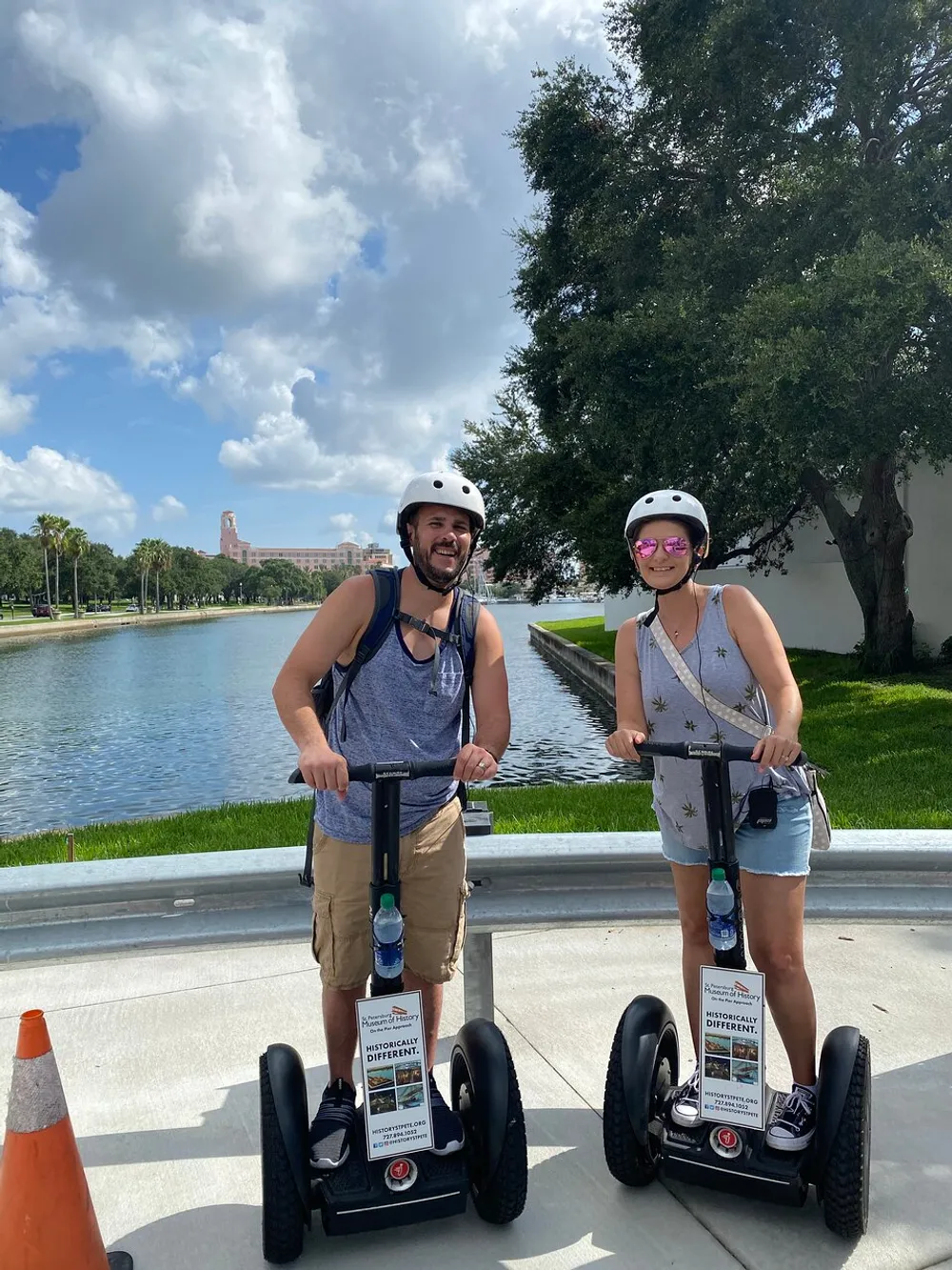 A man and a woman are smiling for the camera while standing on Segways near a waterway with lush greenery and a clear blue sky in the background