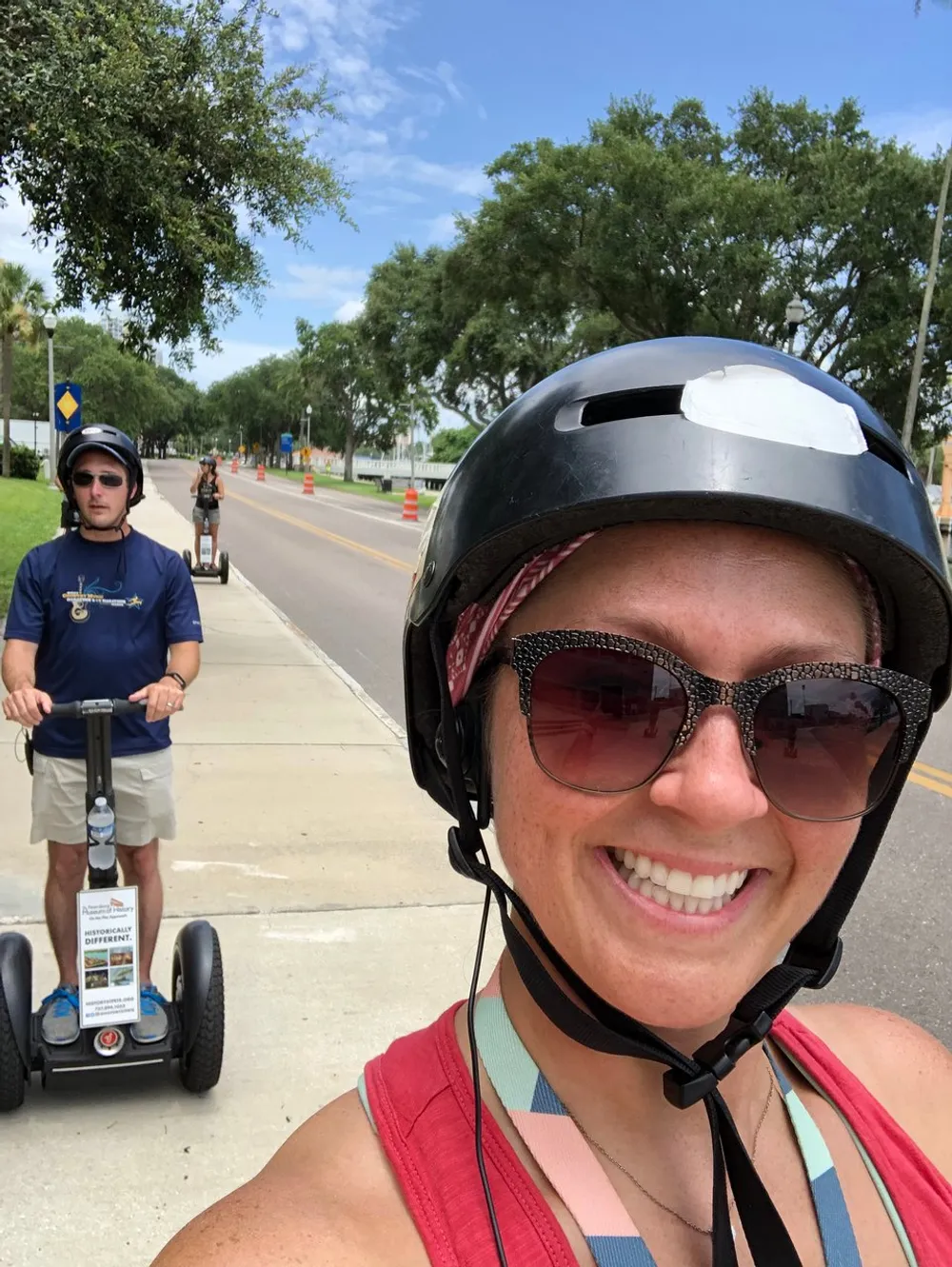 A person is taking a selfie while wearing a helmet smiling with another person riding a Segway in the background