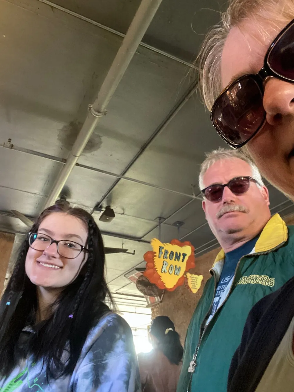 Three people two of whom are wearing sunglasses are posing for a selfie with a smile and there is a sign that reads FRONT ROW in the background