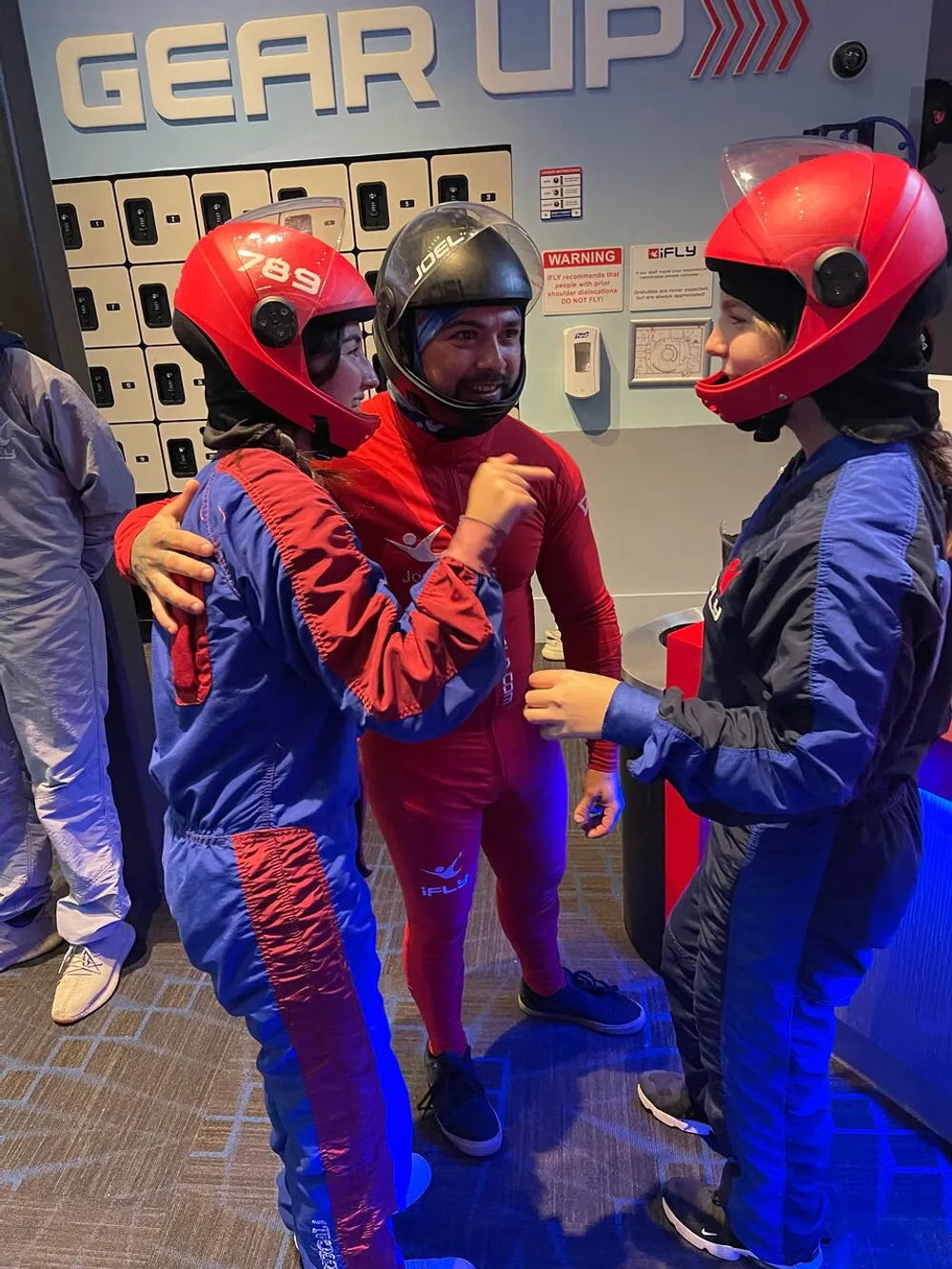 Three individuals wearing flight suits and helmets appear to be in a lively conversation before an indoor skydiving experience with lockers and flight gear instructions visible in the background