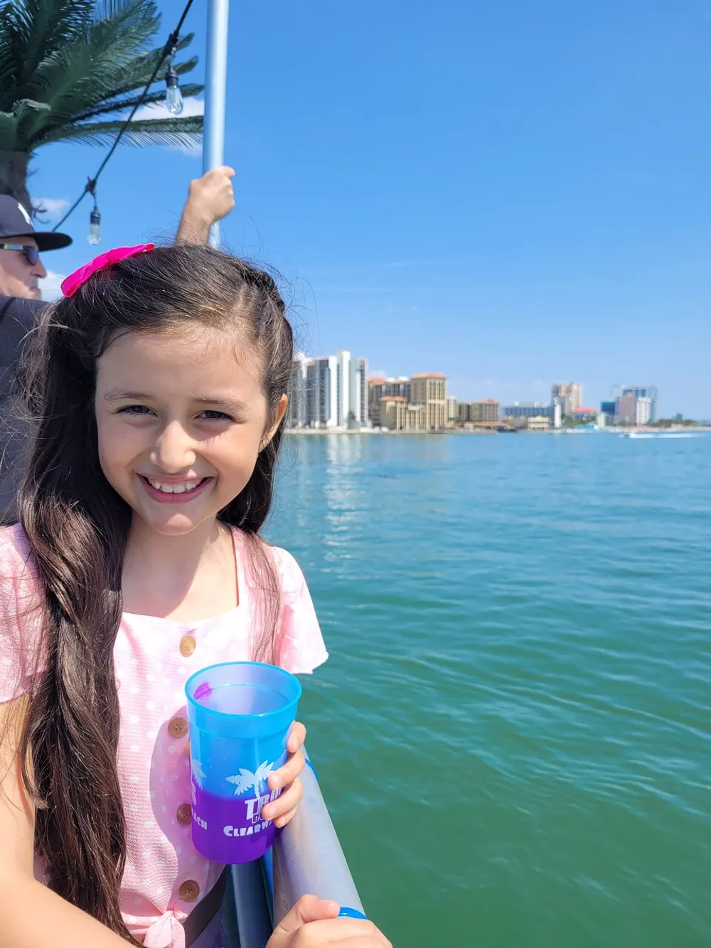 A smiling young girl is holding a blue cup and standing by the railing of a boat with a city skyline in the background