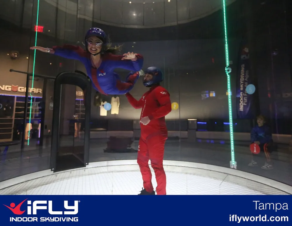 A person is enjoying indoor skydiving in a vertical wind tunnel with an instructor nearby while someone watches from the side