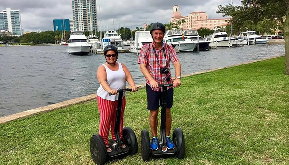 Two people are standing on Segways by a waterfront with boats and buildings in the background