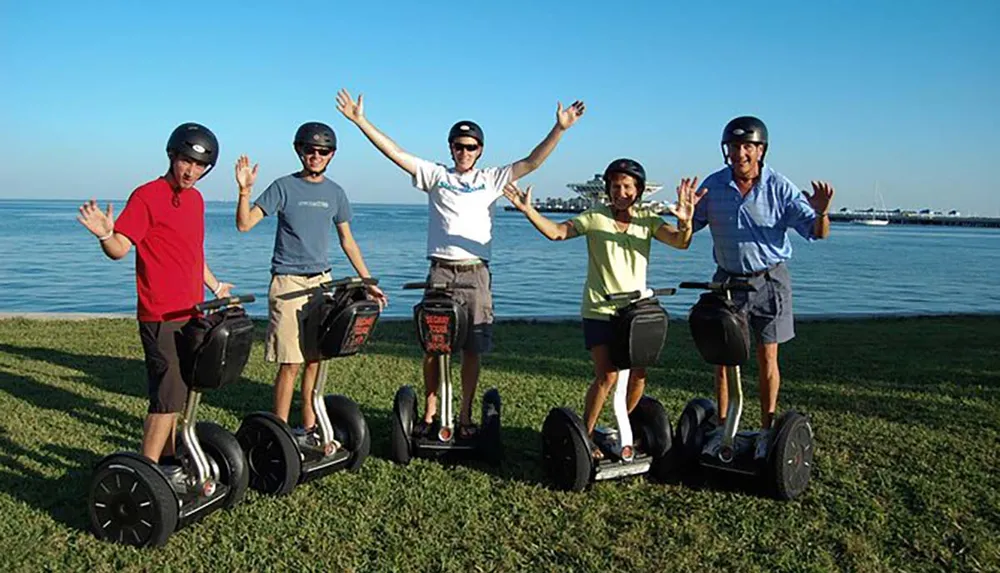 A group of five people wearing helmets are smiling and posing on Segways by the seaside