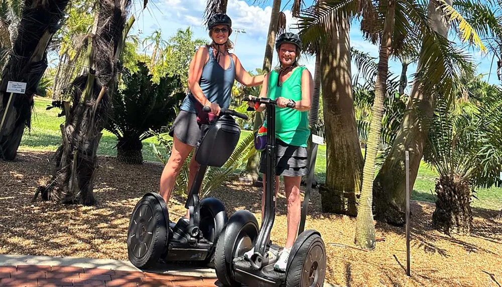 Two people are happily riding Segways outdoors on a sunny day surrounded by lush greenery