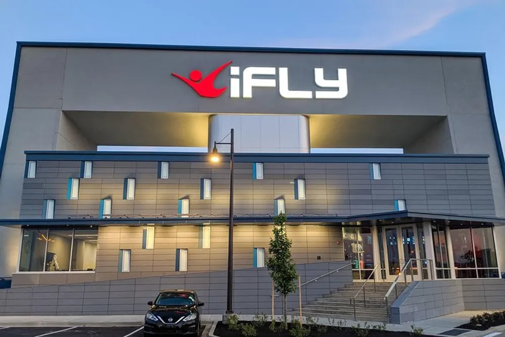 The image shows the modern exterior of an iFLY indoor skydiving facility during dusk or evening with the lit up brand logo prominently displayed above the entrance
