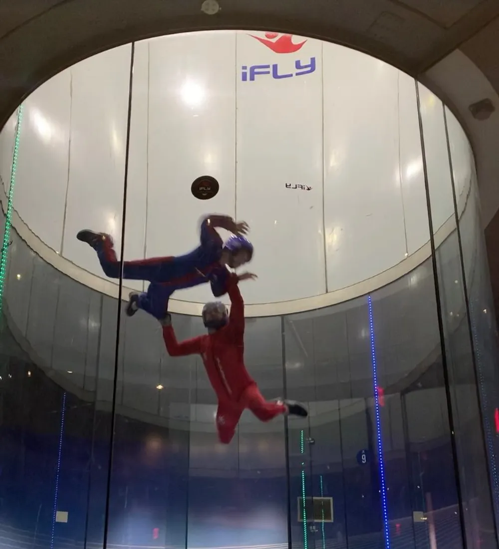 Two people are indoor skydiving in a vertical wind tunnel floating in the air while wearing jumpsuits and helmets