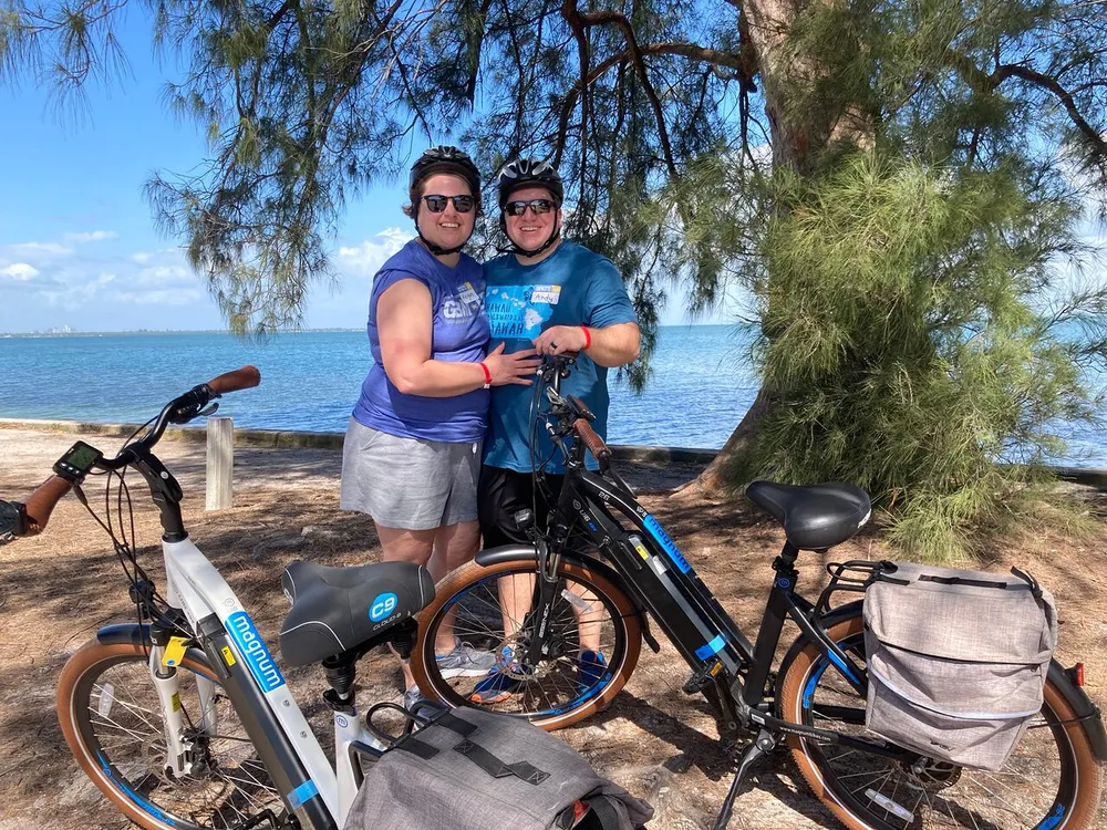 Two happy individuals are posing with their bicycles by a serene beachside with trees in the background
