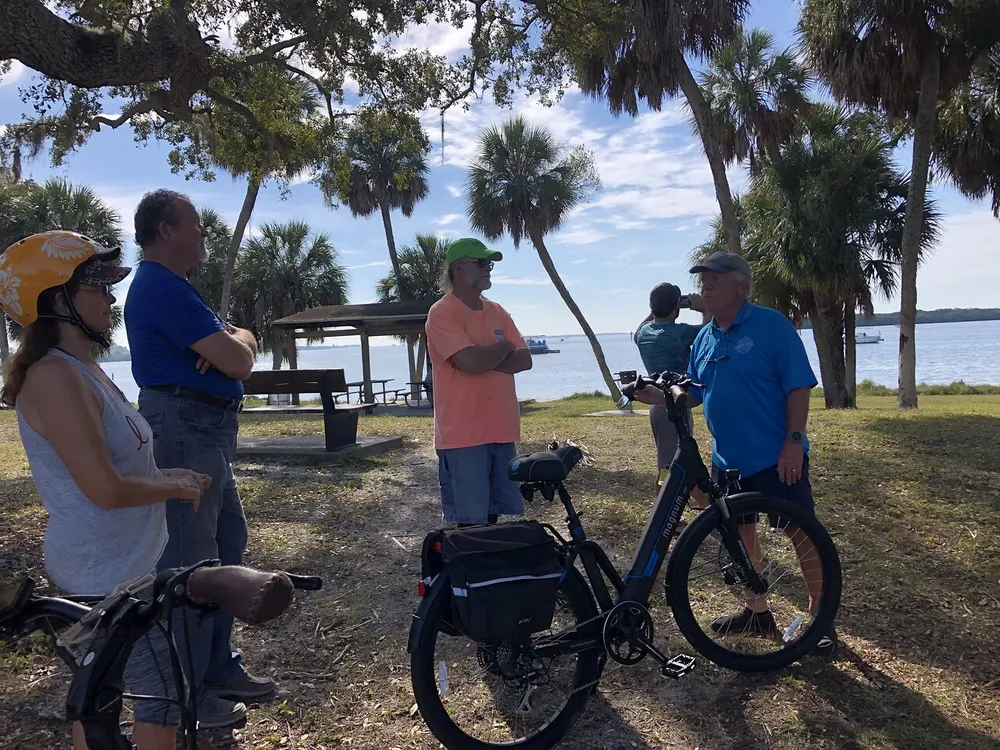 A group of people some with bicycles appear to be conversing in a scenic park with palm trees and a view of the water