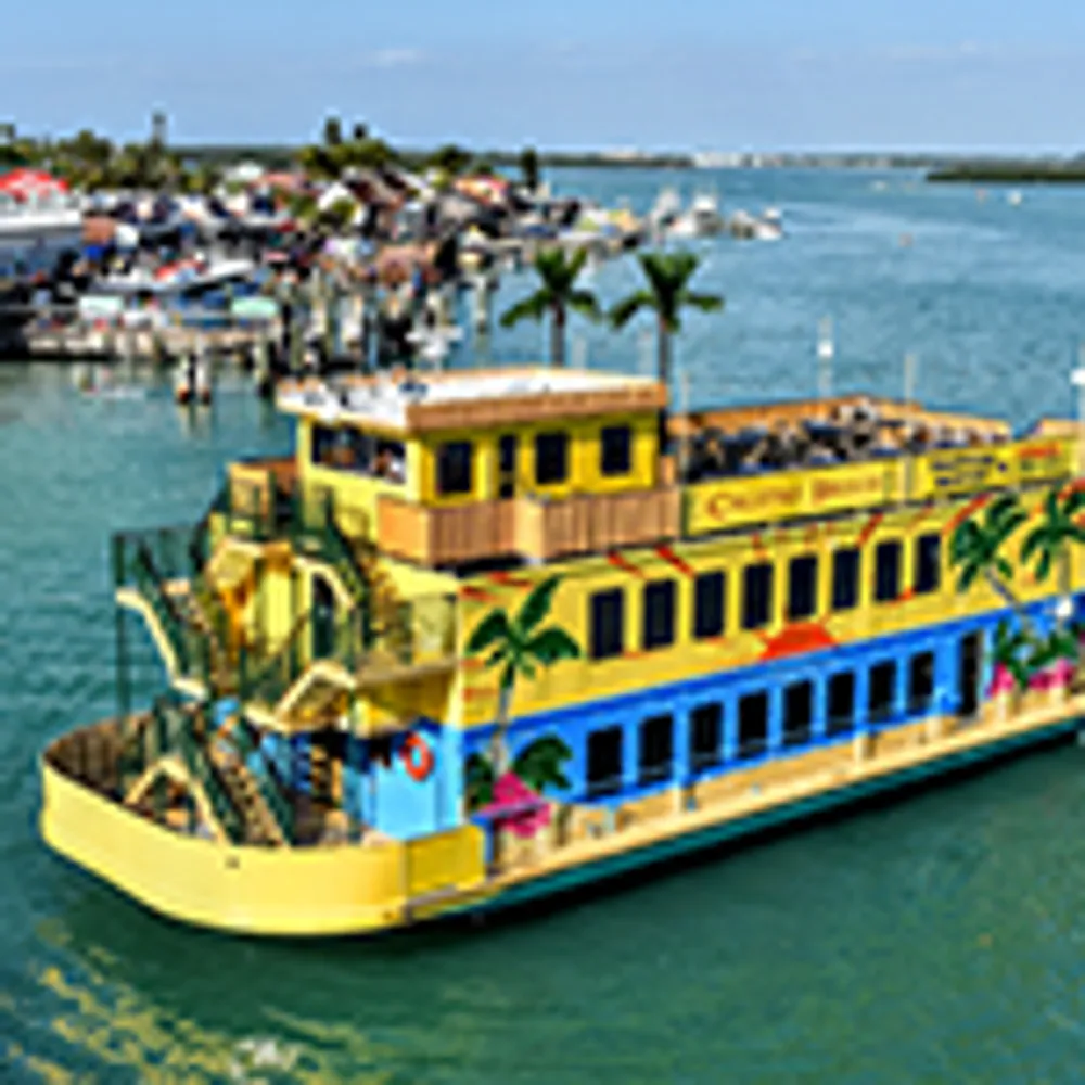 The image shows a colorful palm tree-adorned boat sailing through a calm body of water with a bustling marina in the background