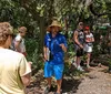 A person in a straw hat and blue outfit appears to be leading an outdoor tour or giving a presentation to a group of attentive listeners in a natural wooded setting
