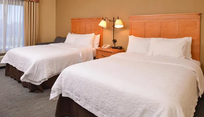 The image shows a neatly arranged hotel room with two large beds a nightstand with a lamp and a window with curtains