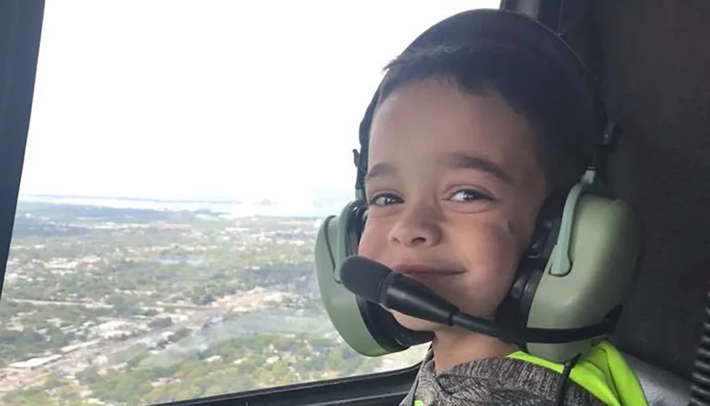A young child wearing aviation headphones is smiling inside a helicopter with a scenic aerial view in the background
