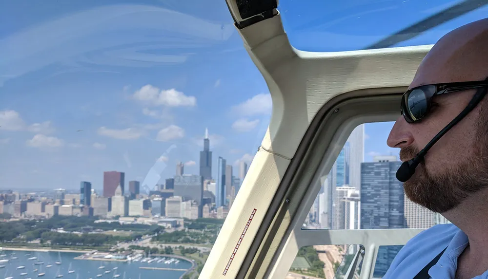 A pilot wearing sunglasses and a headset is looking out from the cockpit of a helicopter with a panoramic view of a cityscape in the background
