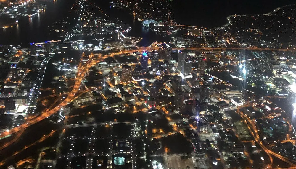 The image shows a nighttime aerial view of a densely built-up urban area illuminated by numerous lights and crisscrossed by glowing streets and highways