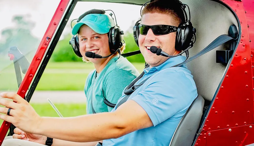 Two smiling people wearing headsets are sitting inside a red helicopter suggesting they might be pilots or passengers preparing for a flight