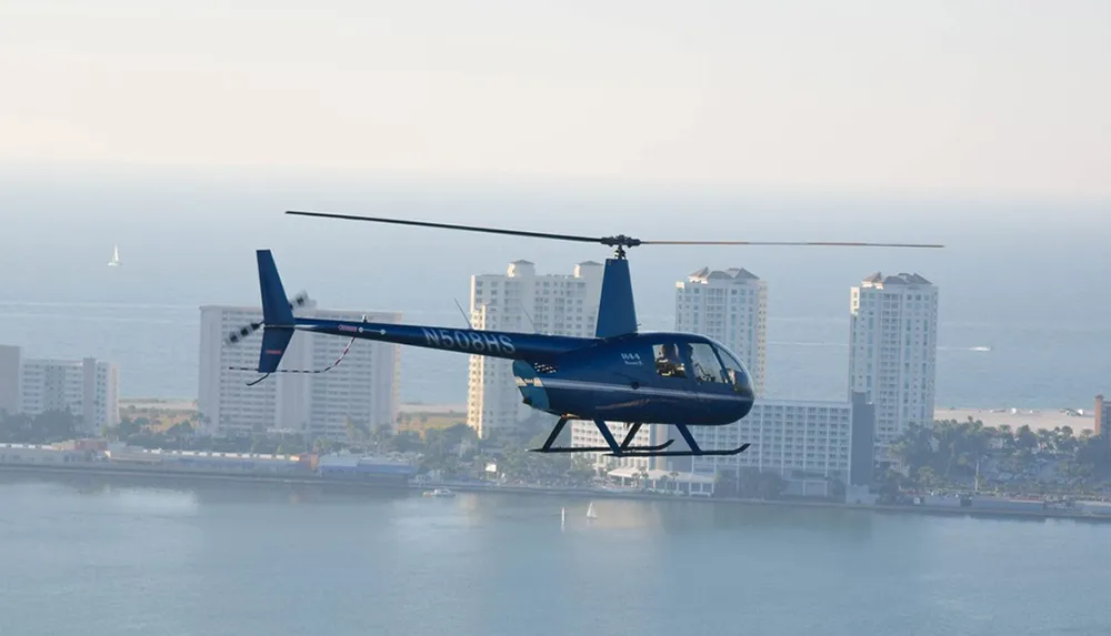 A blue helicopter is flying over a coastal cityscape with high-rise buildings in the background