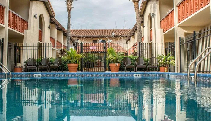 The image captures a tranquil outdoor swimming pool area encircled by a metal fence with patio furniture and potted plants set against a backdrop of a two-story building with a Spanish architectural influence