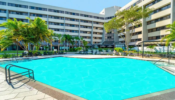 The image shows a large swimming pool with clear blue water surrounded by lounge chairs and palm trees in front of a multi-story hotel building on a sunny day