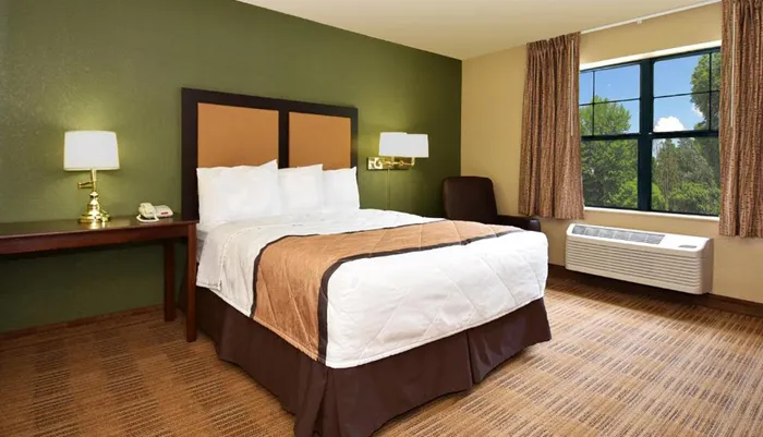 The image shows a neatly arranged hotel room with a queen-sized bed two bedside tables with lamps a window with a view of trees and an air conditioning unit below the window