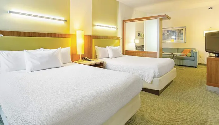 The image shows a modern well-lit hotel room with two double beds a sitting area with a sofa and a television