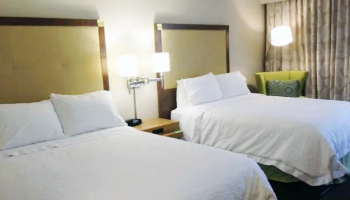 This image shows a tidy hotel room with two made-up beds bedside lamps and a green accent chair