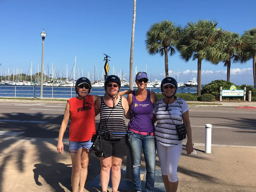 Four people are posing for a photo with helmets on a sunny day near a marina lined with palm trees and boats adjacent to a park sign