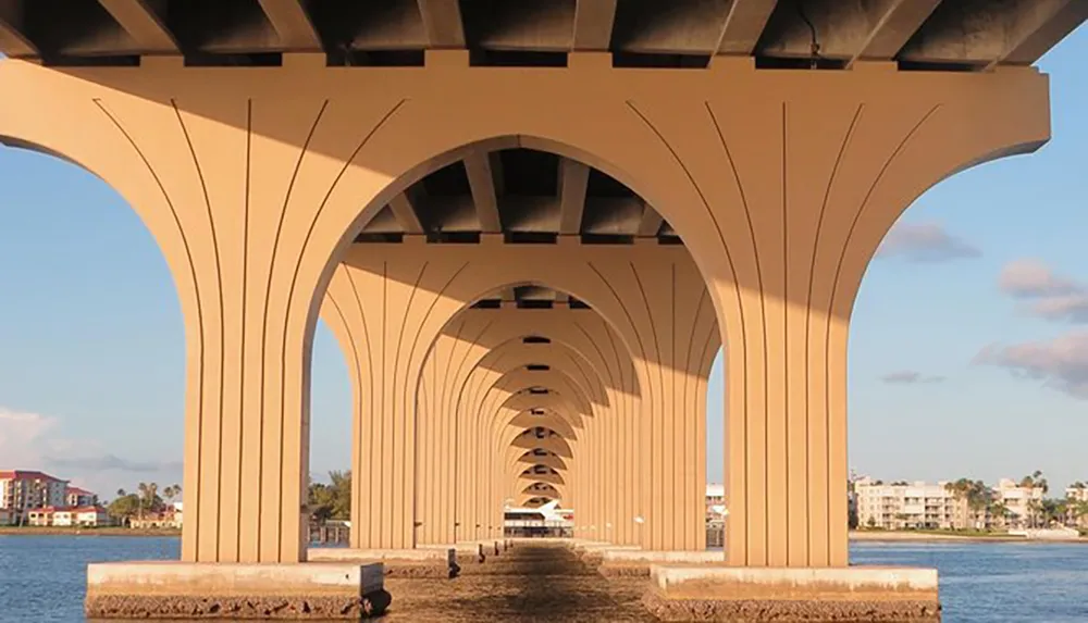 The image shows the underside of a high arched bridge with linear symmetry as it stretches over a body of water with a view of buildings in the distance