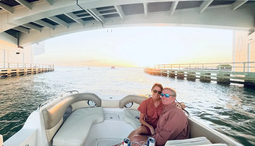Two people are smiling and enjoying a boat ride near a bridge at sunset