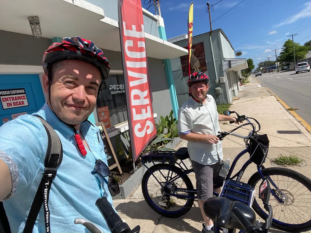 Two individuals wearing bike helmets pose for a selfie on a sunny day one holding the phone and the other standing by an electric bicycle with a sign for electric bike service in the background