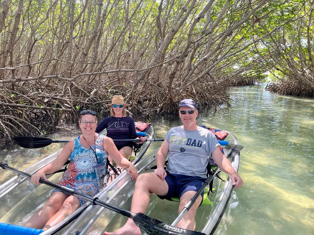 Three people are enjoying a clear kayak tour through a mangrove tunnel smiling and looking relaxed in the serene natural setting